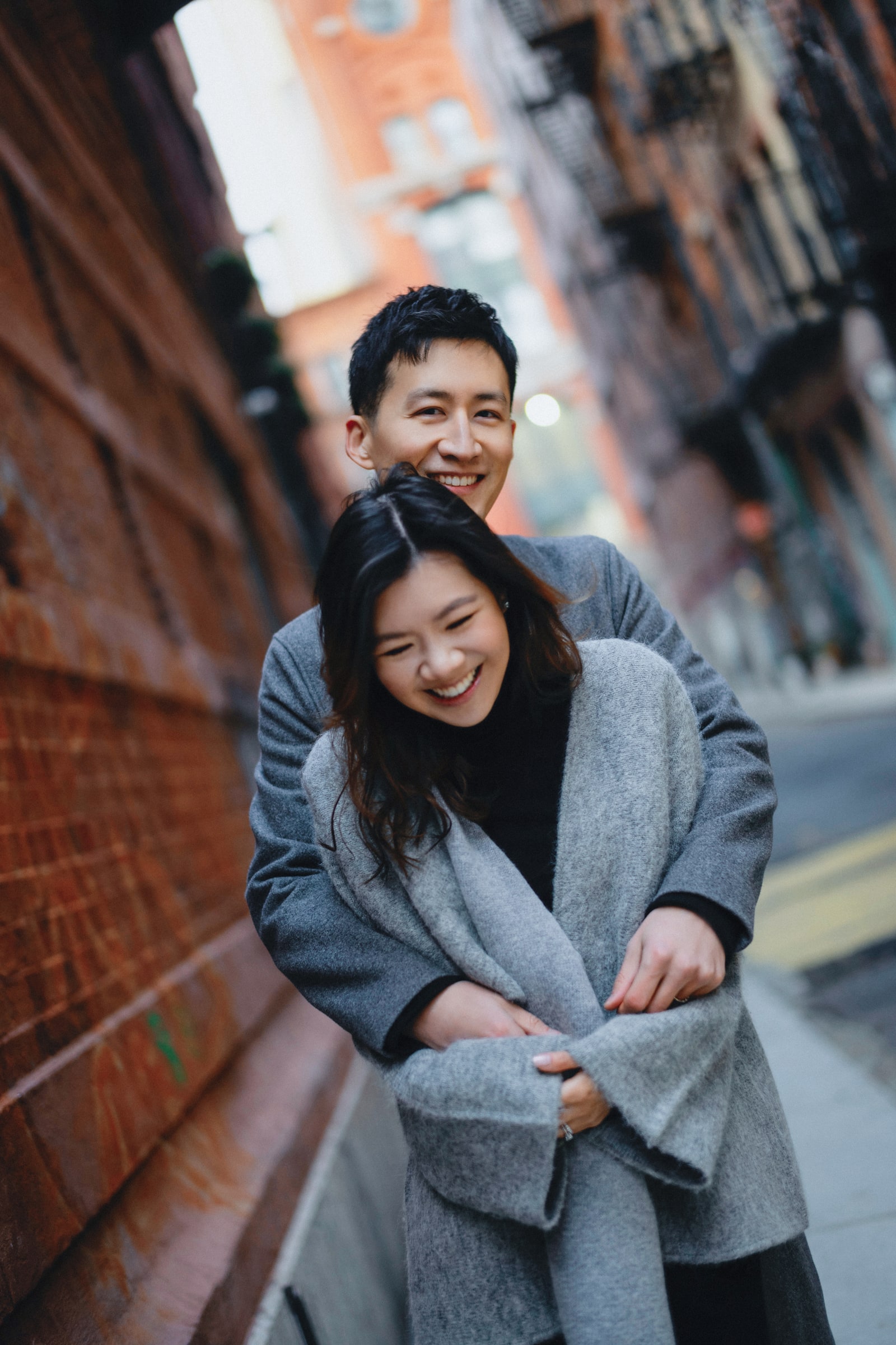 A laughing couple in grey coats embracing in a brick-lined alley