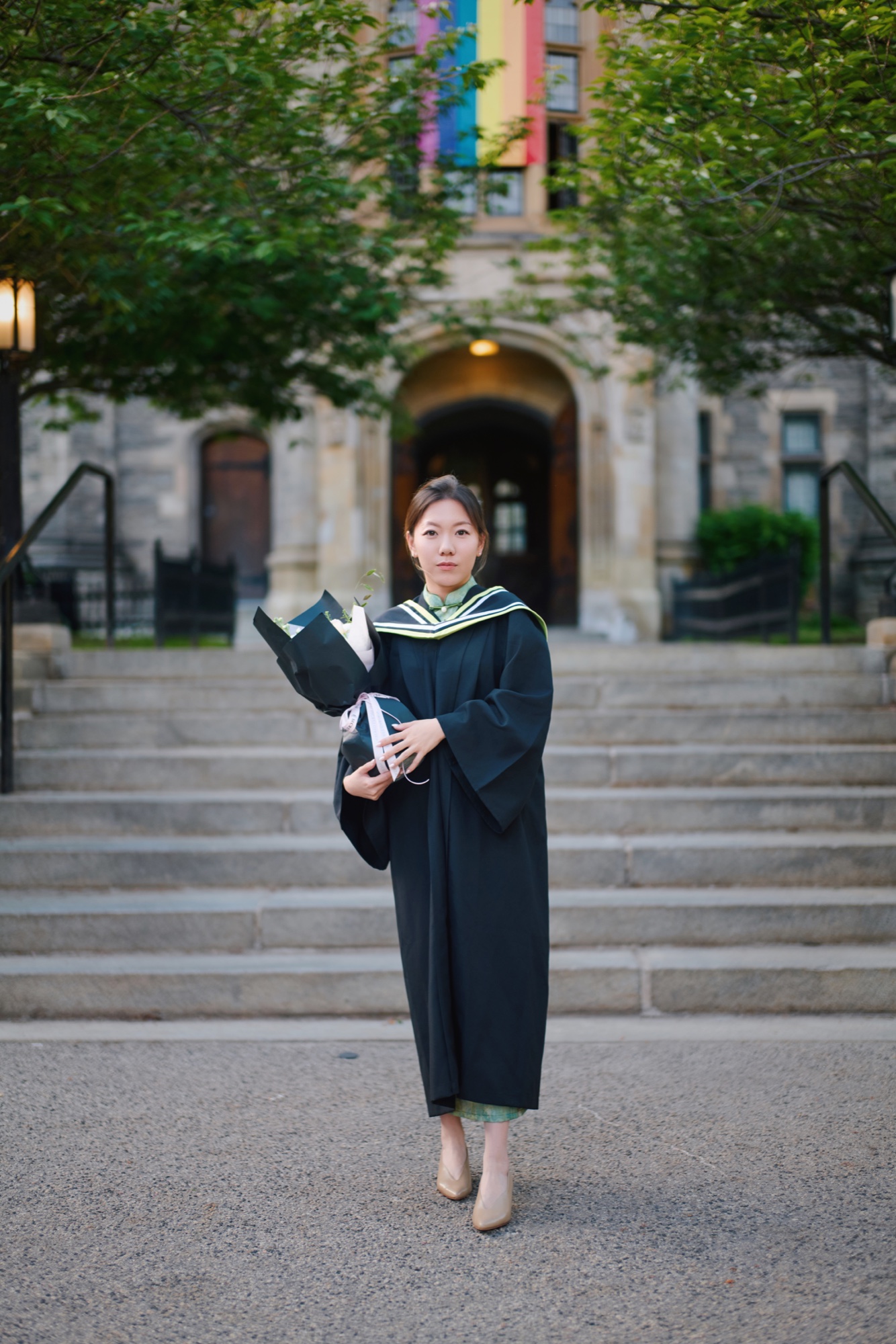 A graduate holding a bouquet in front of tree-lined campus steps