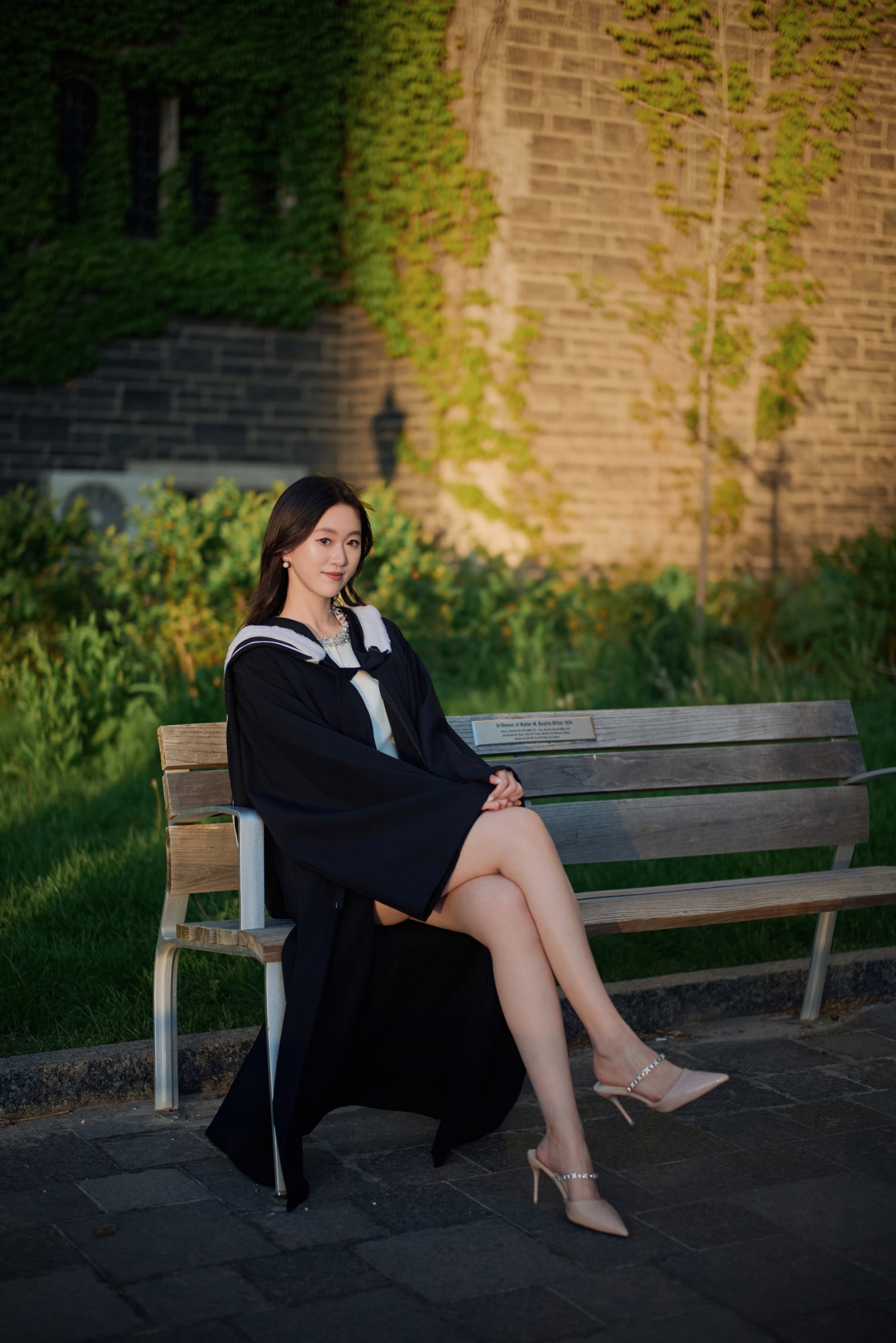 A graduate seated on a wooden bench beside an ivy-covered wall in late afternoon light