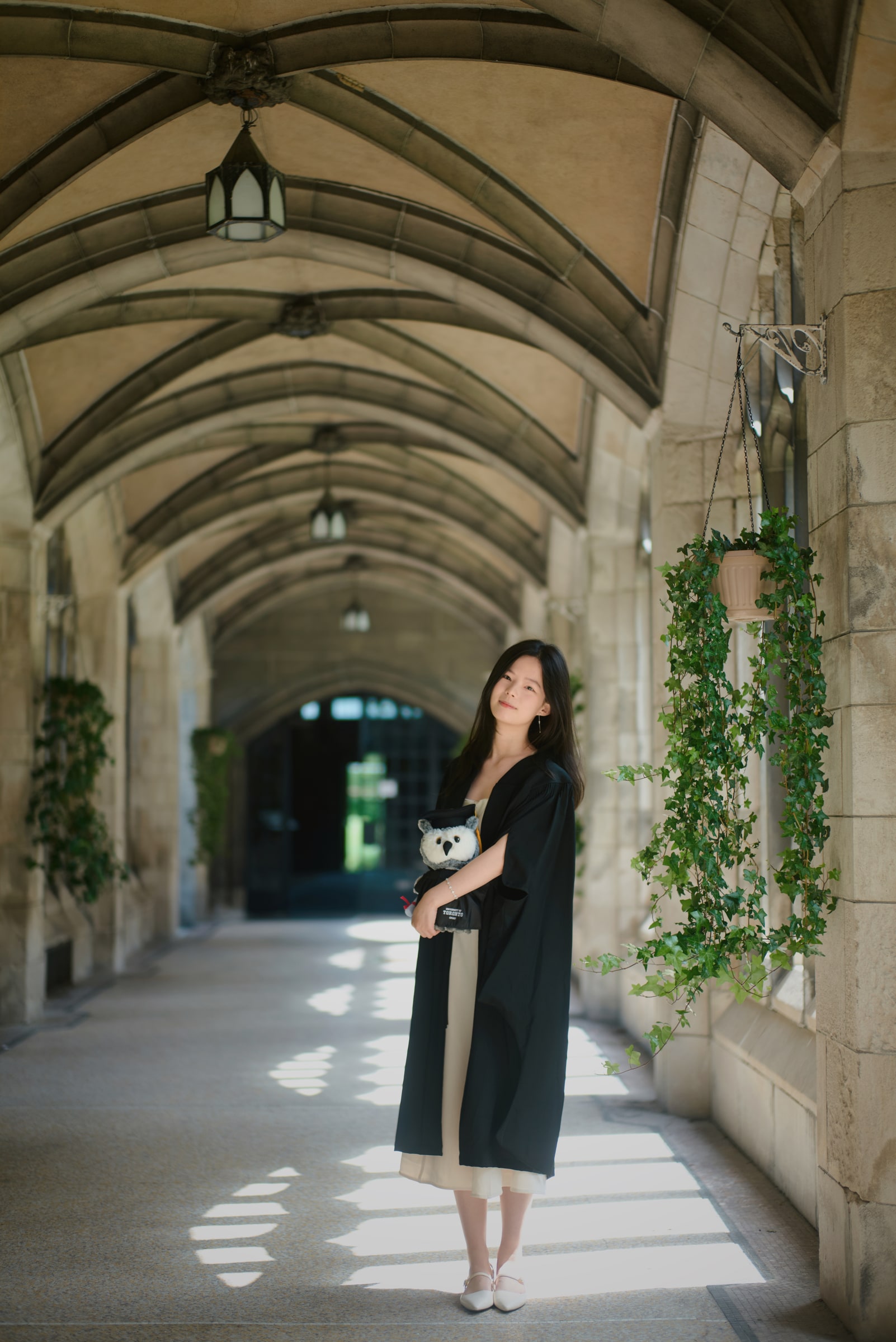 A graduate holding a plush owl beneath a vaulted stone walkway with hanging ivy