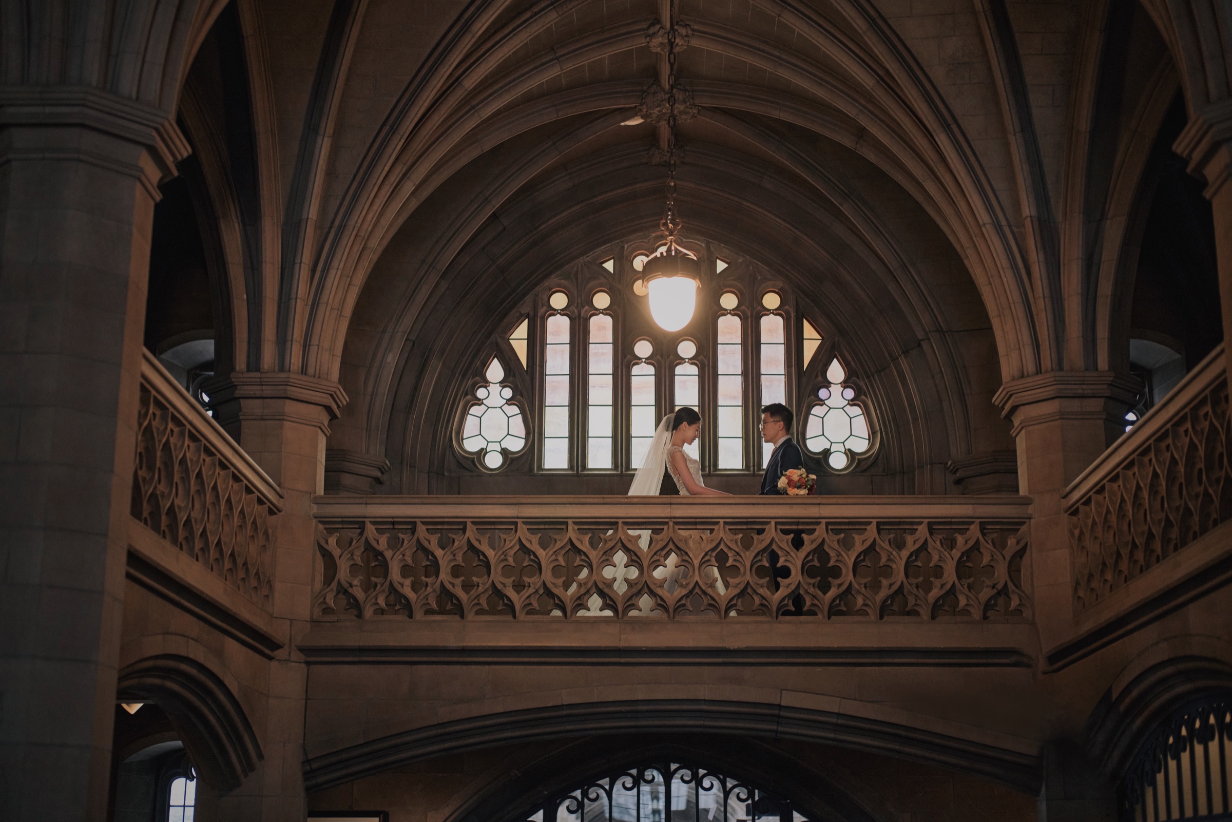 A wedding couple beneath vaulted stone arches, with a bouquet beside a tall window